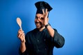 Young cooker man with beard wearing uniform holding wooden spoon over blue background with happy face smiling doing ok sign with Royalty Free Stock Photo