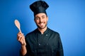 Young cooker man with beard wearing uniform holding wooden spoon over blue background with a happy face standing and smiling with Royalty Free Stock Photo