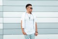 Young cool American man hipster in sunglasses in blue trendy jeans in a fashionable white t-shirt is poses in the city near a Royalty Free Stock Photo