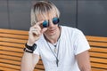 Young cool hipster man blond in a trendy white t-shirt with vintage amulets around neck sits on a yellow wooden bench and Royalty Free Stock Photo