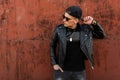 Young cool hipster man in stylish T-shirt in trendy black baseball cap with dark glasses in a leather trendy black jacket Royalty Free Stock Photo