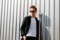 Young cool hipster man with a trendy hairstyle in elegant clothes in fashionable dark sunglasses poses near a silver metal wall