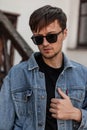 Young cool man straightens denim fashionable blue jacket. Attractive urban guy hipster in trendy sunglasses Royalty Free Stock Photo