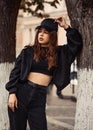 Young cool teen hipster woman looking serious near the tree on the street background in black style clothing, jeans, jacket and