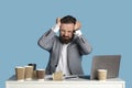 Young corporate worker whining or crying, grabbing his head in despair, sitting at office desk on blue studio background Royalty Free Stock Photo