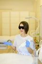 Young cosmetologist wearing special purpose glasses and latex gloves sitting near permanent makeup device.