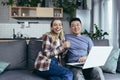 Young couple Asian man and woman at home sitting on sofa together using laptop, happy and smiling multiracial family looking at Royalty Free Stock Photo