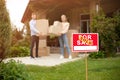 Young couple with boxes in front of their new house on moving day, focus on FOR SALE sign. Blank space Royalty Free Stock Photo