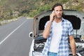 Man standing beside car with open hood holding smartphone speaking on mountain road near guardrail Royalty Free Stock Photo