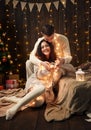 Young couple is in christmas lights and decoration, dressed in white, fir tree on dark wooden background, winter holiday concept Royalty Free Stock Photo