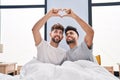 Young couple doing heart symbol with hands sitting on bed at bedrooom