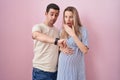 Young couple expecting a baby standing over pink background looking at the watch time worried, afraid of getting late