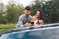 Young couple is looking at something cheerful in the smartphone, standing leaning on the hood of car.