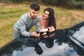 Young couple is looking at something cheerful in the smartphone, standing leaning on the hood of car.