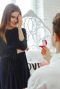 Young couple in love at home. Smiling man holding red box with marriage engagement ring got up on his knee and make proposal to