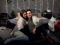 Young couple in love,sitting near bed with christmas lights behind, embracing, kissing. Brunette man and woman, wearing grey