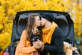 A young couple in love is sitting on the open trunk of a black car with their little dog in the autumn forest. Lovers kiss, and