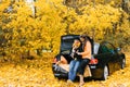 A young couple in love is sitting on the open trunk of a black car with their little dog in the autumn forest. Lovers look at Royalty Free Stock Photo