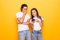 Young couple man and woman in basic t-shirts holding and peeking at cell phones isolated over yellow background Royalty Free Stock Photo