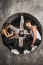 A young couple a man and a woman with a playful mood, fashionable and sporty look sit near an industrial fan in the factory with a