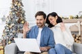 Young couple man and woman sitting on sofa in front of laptop, talking on video call, waving and greeting at camera