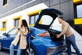 Young couple man and woman traveling by electric car having stop at charging station. The man taking a suitcase from the trunk. Royalty Free Stock Photo