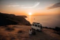 young couple sits on a hill above the sea next to their travel minivan vehicle at sunset, neural network generated