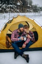 Young couple sits in tent and drinks hot tea during winter hike.