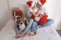 young couple sitting on bed with christmas santa hats having fun with their dog. Presents on bed. Home, indoors. Xmas concept Royalty Free Stock Photo