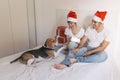 young couple sitting on bed with christmas santa hats having fun with their dog. Presents on bed. Home, indoors. Xmas concept Royalty Free Stock Photo