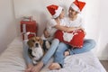 young couple sitting on bed with christmas santa hats having fun with their dog. Presents on bed. Home, indoors. Xmas concept Royalty Free Stock Photo