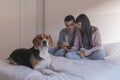 young couple sitting on bed, using mobile phone and having fun. cute beagle dog besides. Breakfast time. Home, indoors Royalty Free Stock Photo