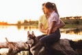 A young couple is sitting by the lake at sunset with a Husky breed dog.