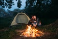 A young couple are sitting near a tent in the forest and making a bonfire in the evening. The branches of the tree are Royalty Free Stock Photo