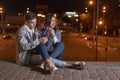 Young couple is sitting on the pavement embracing on evening city background with garland in their hands. Night city lights Royalty Free Stock Photo