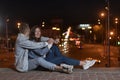 Young couple sitting on the pavement smiling and hugging each other against the backdrop of the lights of the night city. Date and Royalty Free Stock Photo