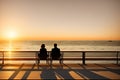 Young couple sitting on the pier by the lake during sunset. A man and a woman, embracing, look at the sun, generative ai Royalty Free Stock Photo