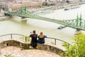 A young couple sitting at a viewpoint up high taking pictures of Liberty bridge over the Danube river, Budapest