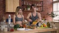 Young couple in sports clothes standing behind kitchen table preparing on lunc fresh salad with vegetables in the