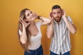Young couple standing over yellow background trying to hear both hands on ear gesture, curious for gossip