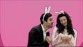 Young couple standing standing on pink background. During this time, they are dressed in rabble ears. Looking at each