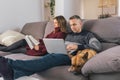 Young couple and their dog enjoying leisure time and relaxing on the couch at home, reading a book and using a laptop. Couple life Royalty Free Stock Photo