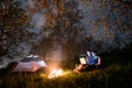 Couple tourists using laptop in the camping at night. Pair sitting near campfire and tent under trees and night sky Royalty Free Stock Photo