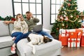 Young couple using laptop sitting by christmas tree doing ok gesture with hand smiling, eye looking through fingers with happy Royalty Free Stock Photo
