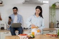 Young couple using laptop and smartphone in the kitchen, smiling man working online at home while his wife cooking with phone apps Royalty Free Stock Photo