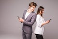 Young couple is using smart phones and smiling while standing back to back on a gray background. Man look at woman. Royalty Free Stock Photo
