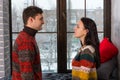 Young couple in warm knitted sweaters looking at each other while standing near windowsill Royalty Free Stock Photo