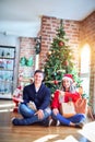 Young couple wearing santa claus hat sitting on the floor around christmas tree at home amazed and smiling to the camera while Royalty Free Stock Photo