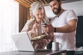 Young couple, woman in bathrobe and towel on her head, stands in kitchen near table with laptop. Freelancers work home.