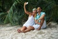 The young couple, the woman and the man sit on the beach and will take the selfies dressed in bright clothes and in sunglasses.